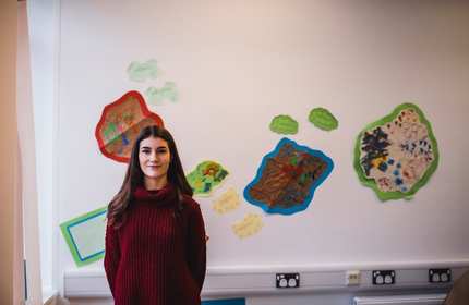 A person with long dark hair wearing a red sweater stands in a classroom setting. The background shows colorful artwork displayed on the wall.