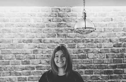 A black and white image of a person smiling, standing in front of a brick wall. A pendant light hangs above their head.