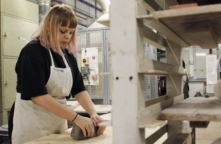 A person wearing an apron moulds a large piece of clay on a table