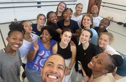 A diverse group of 15 people, mostly women, are smiling and posing for a selfie in a dance studio. They are casually dressed and appear joyful, with some making peace signs. The room has white walls and ballet barres.