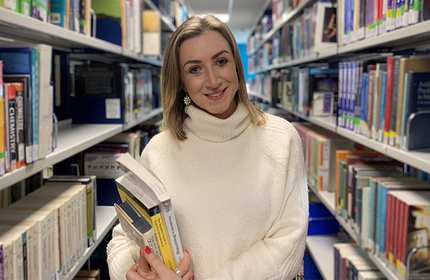 A person wearing a white sweater stands in a library aisle, holding a stack of books. Shelves filled with books line both sides of the aisle. The person smiles at the camera.