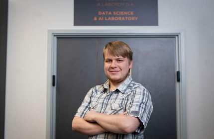 A person stands with arms folded in front of a door. Above the door is a sign that reads Data Science and AI Laboratory.