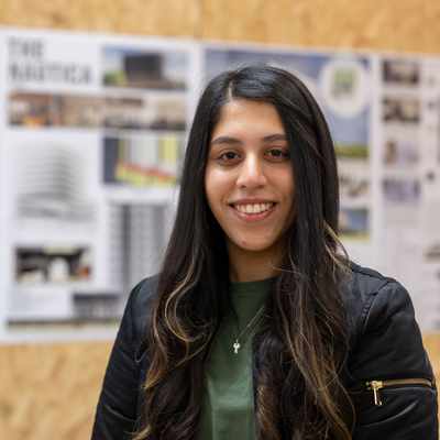 A person with long dark hair and a green shirt smiles at the camera. In the background, there are architectural design boards displayed on a cork wall.