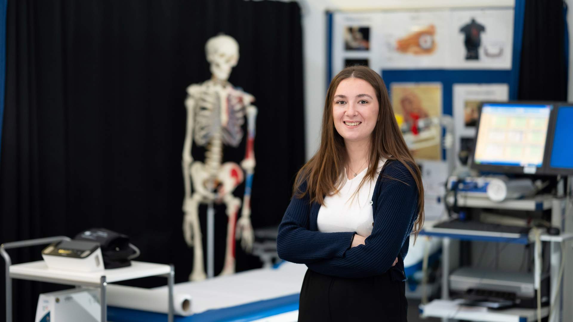 A person in a white lab coat stands smiling in a bright, modern laboratory with blue chairs and yellow safety containers on the tables. The room is well-lit with numerous ceiling lights.