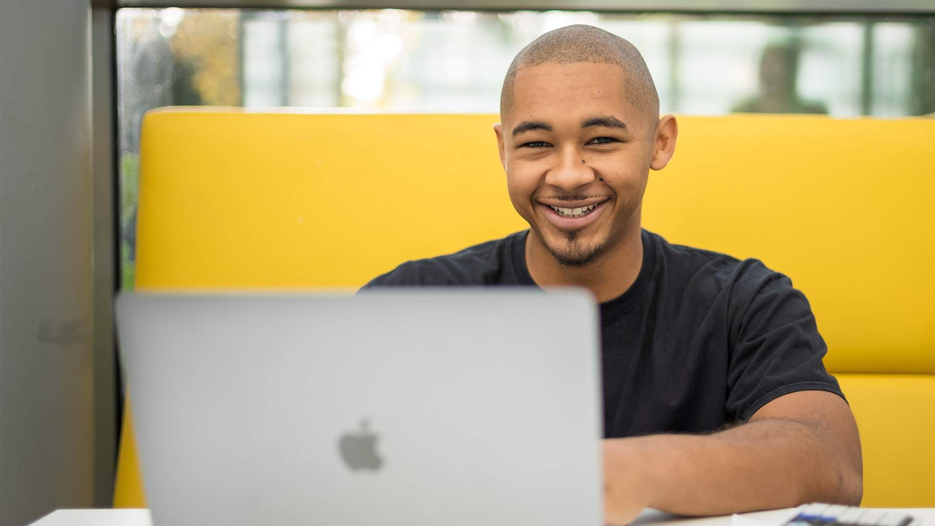 A person is smiling while sitting at a table with a laptop in front of them. They are in a bright, modern setting, with a yellow cushioned seating area behind them.