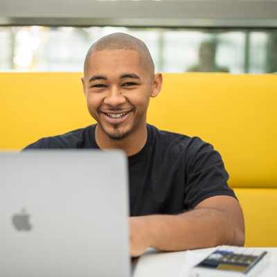 A person is smiling while sitting at a table with a laptop in front of them. They are in a bright, modern setting, with a yellow cushioned seating area behind them.
