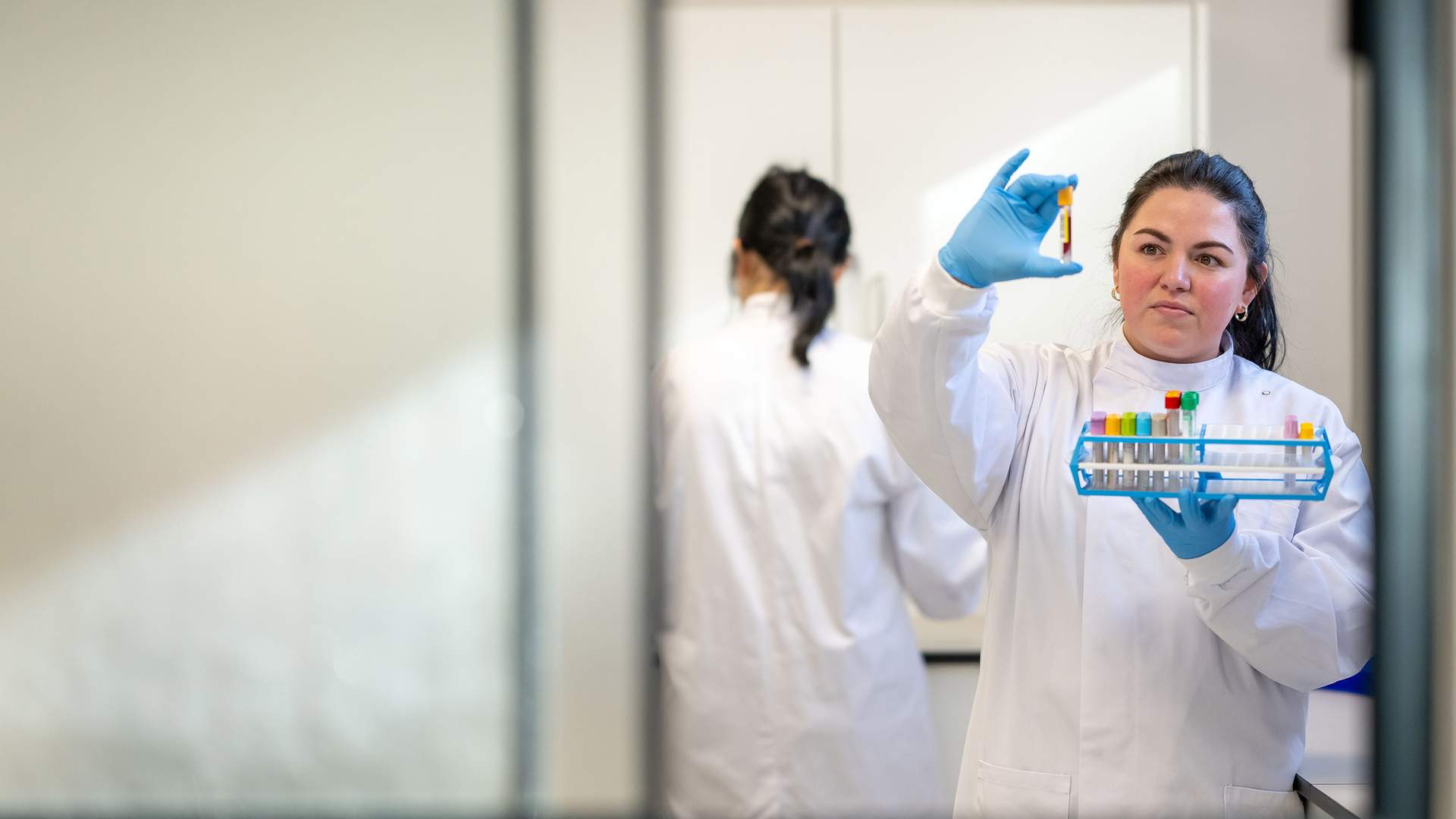 A female scientist in a white lab coat and blue gloves examines a test tube held up to the light. Another person in a lab coat works in the background. Various test tubes are organized in a rack. The scene is viewed through a glass window.