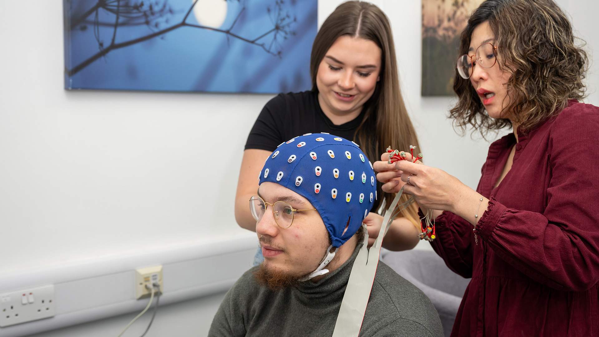 Two people assist a seated person with an EEG cap. One adjusts the cables on the cap, while the other looks on. The room has a calm atmosphere with artwork on the walls.