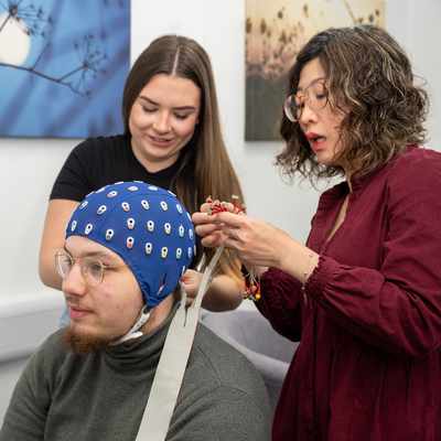 Two people assist a seated person with an EEG cap. One adjusts the cables on the cap, while the other looks on. The room has a calm atmosphere with artwork on the walls.