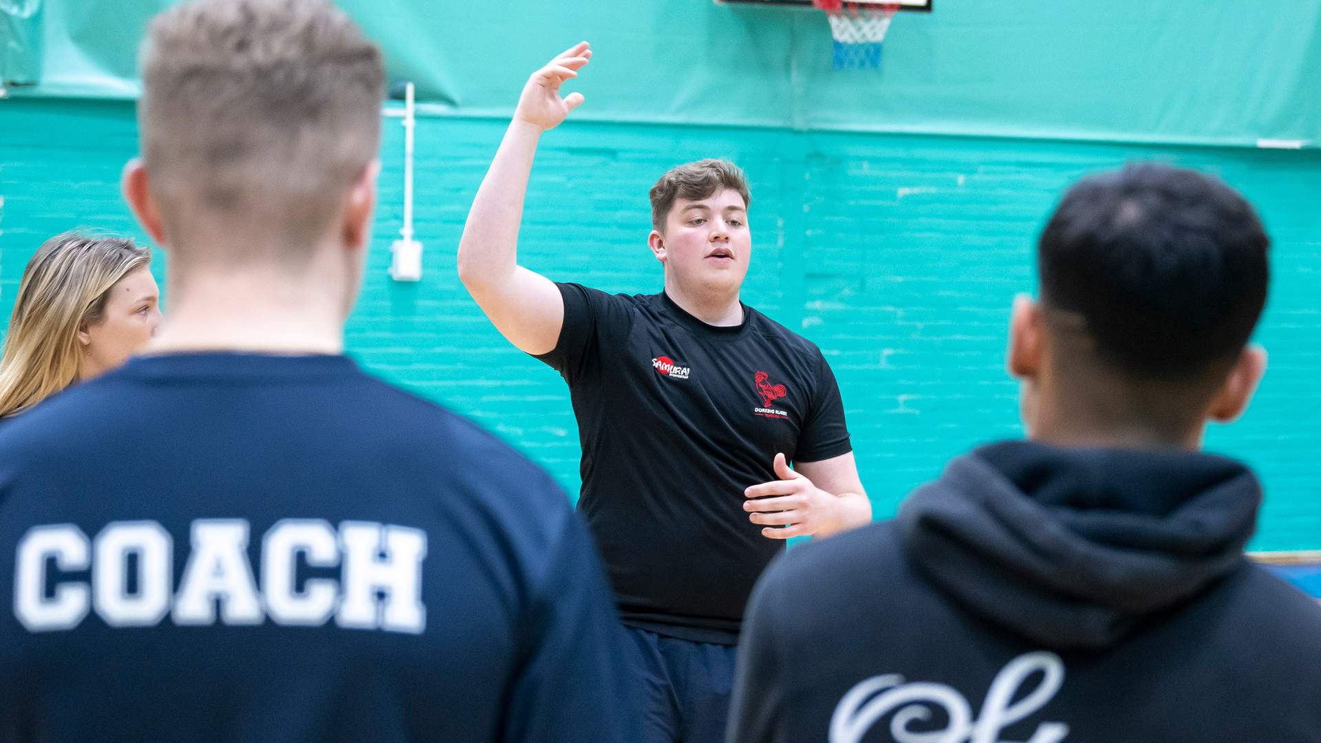 A person stands in a gym, gesturing with one hand raised, facing a group. The back of one person is visible, wearing a shirt labeled Coach. A basketball hoop is in the background. The setting is casual and suggests a team gathering or discussion.