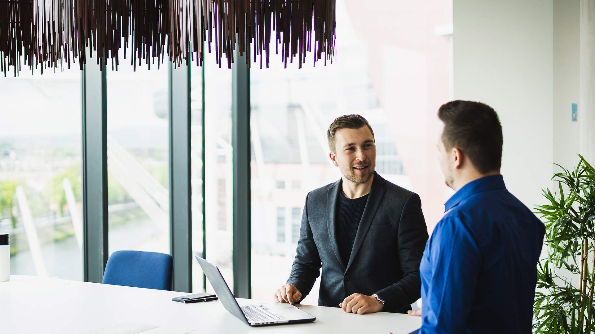 Two men are seated at a white table in a modern office with large windows. One is using a laptop, and they appear to be having a conversation. Natural light fills the room, and theres a potted plant nearby.