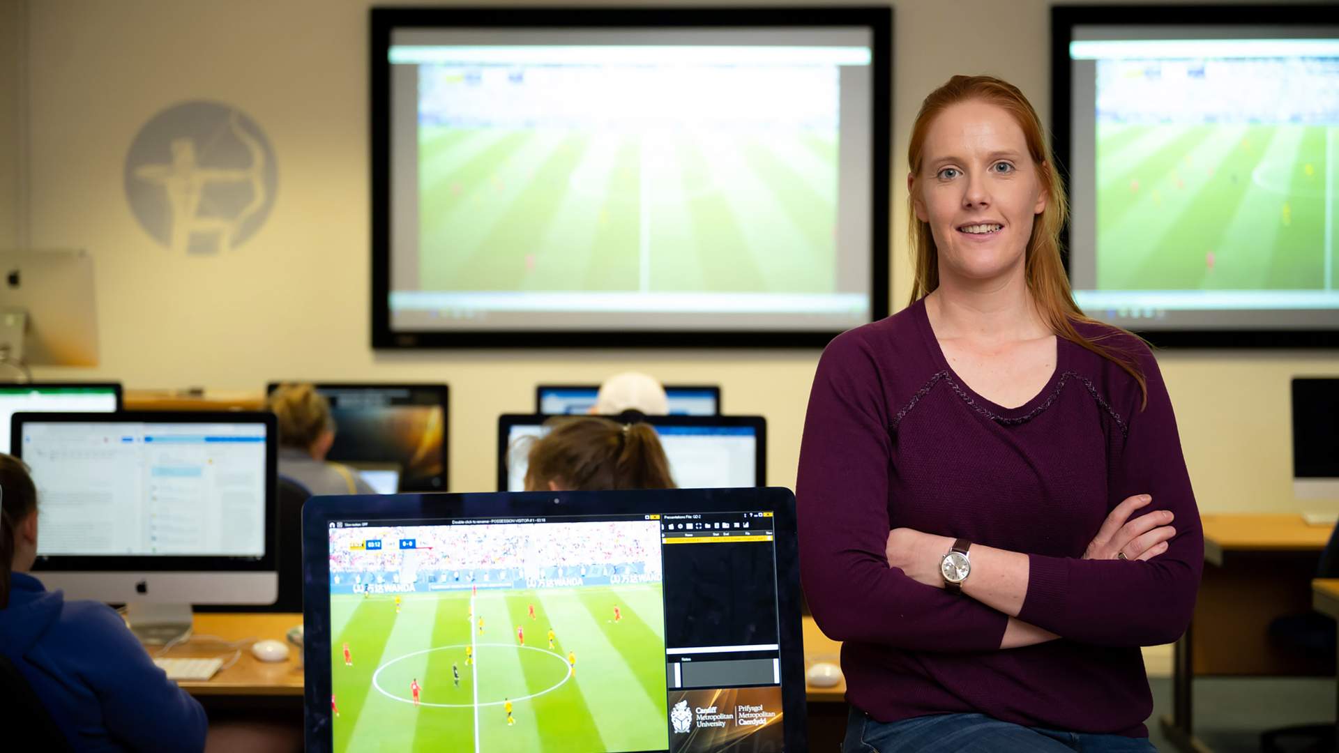 A woman with long red hair, wearing a purple sweater, stands confidently with arms folded in a classroom. People are working at computers, and a soccer game is projected on two large screens in the background.