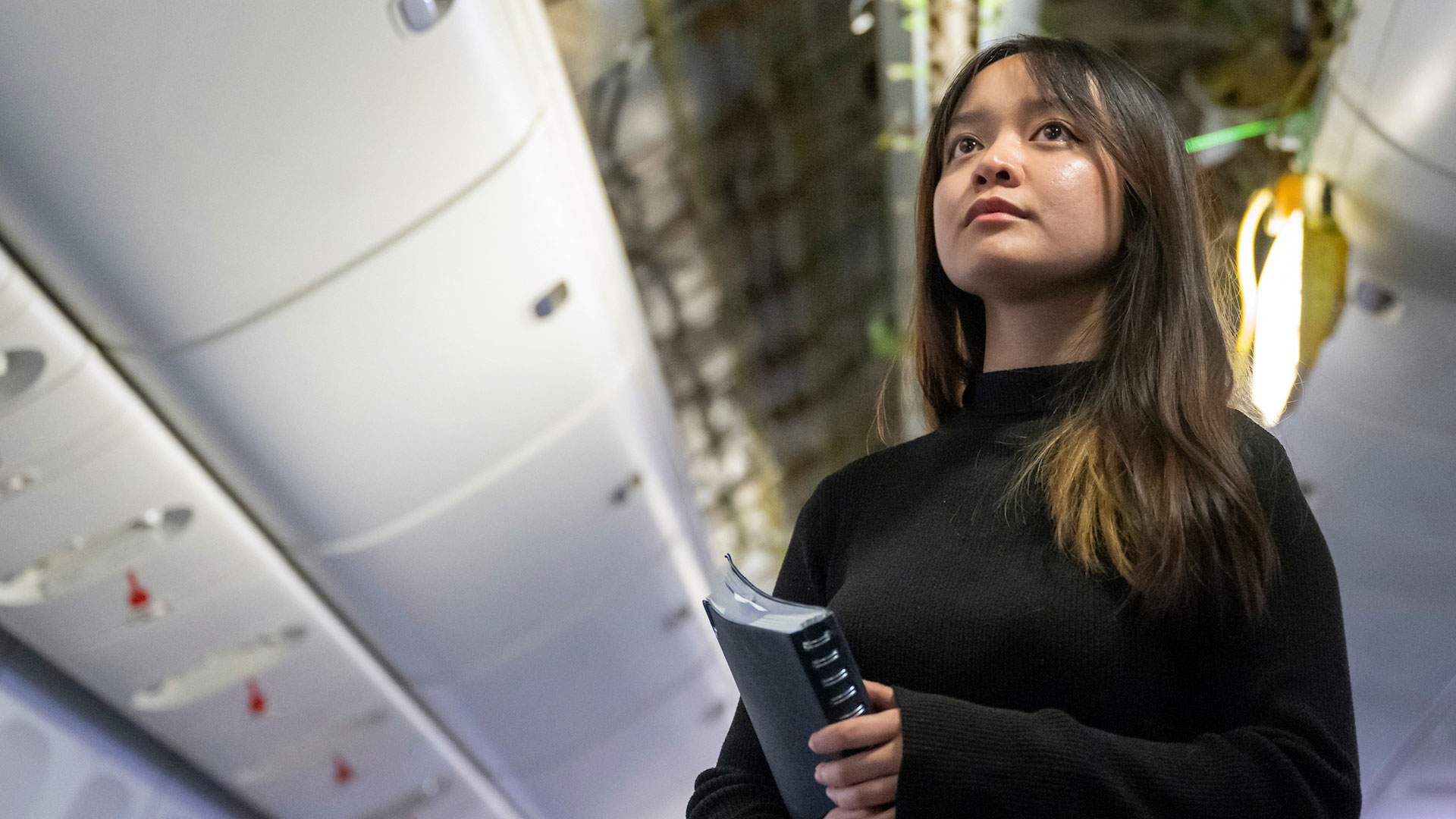 A woman stands inside an airplane, holding a ring binder, inspecting the wiring above.