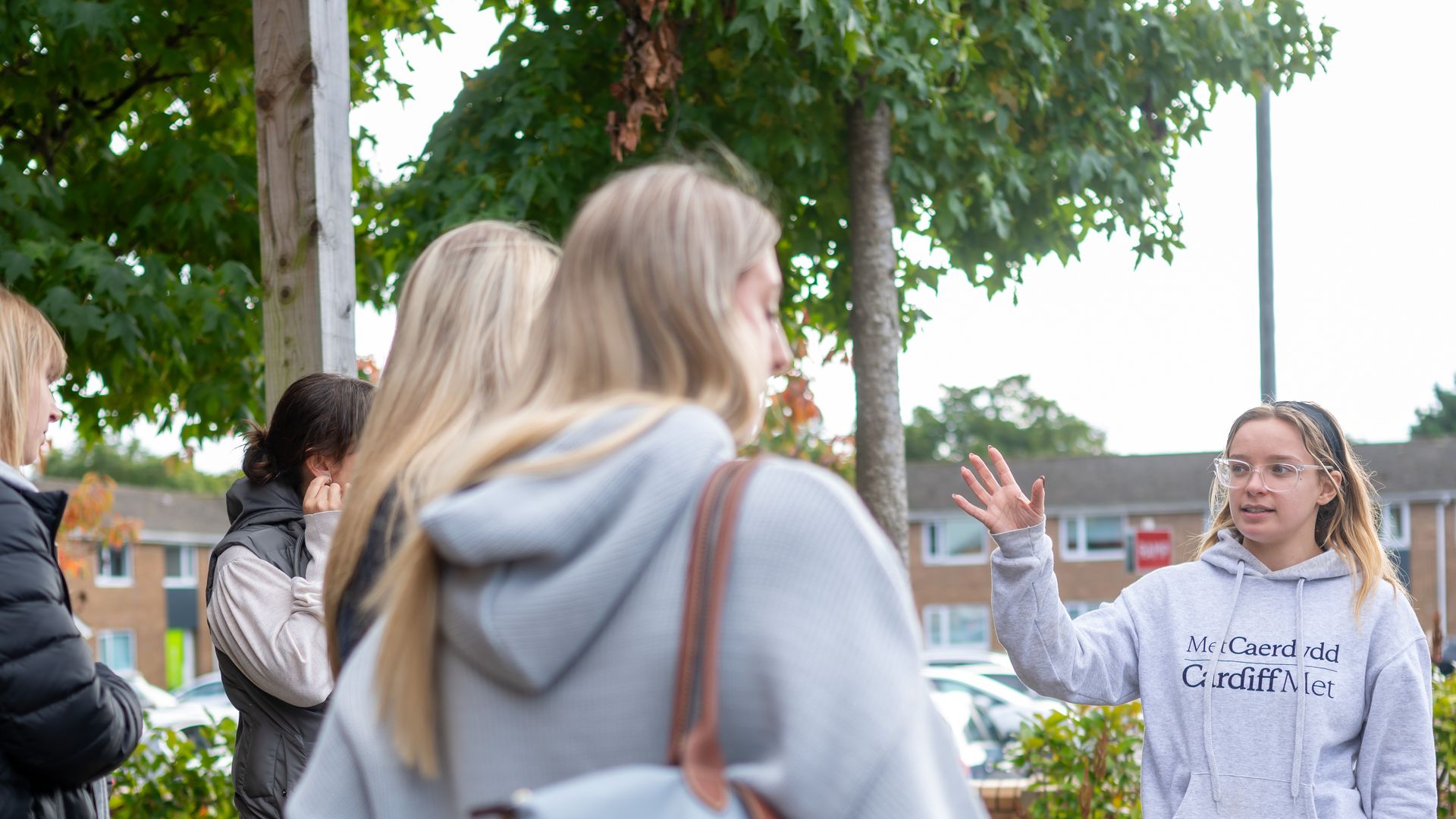 Young woman in grey hoodie and jeans gestures with hand while giving a campus tour to a group of visitors