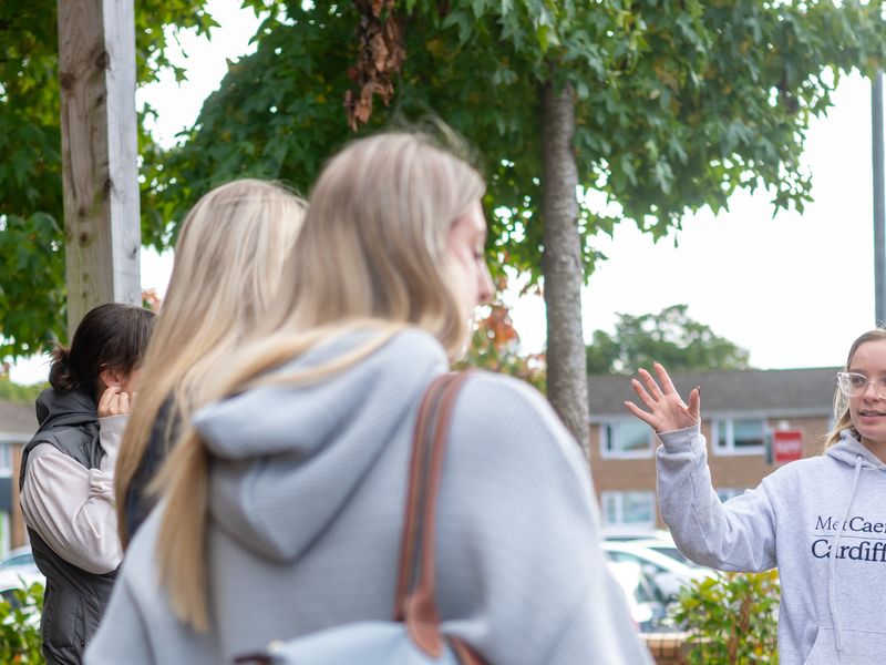 Young woman in grey hoodie and jeans gestures with hand while giving a campus tour to a group of visitors