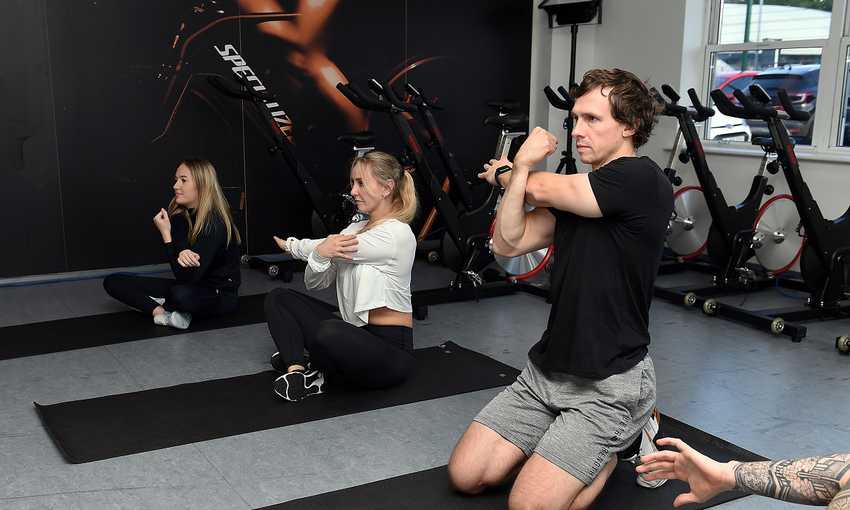A group of people complete arm stretches while sat on their own exercise mats in a dance studio