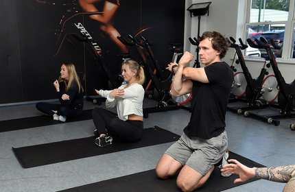 A group of people complete arm stretches while sat on their own exercise mats in a dance studio