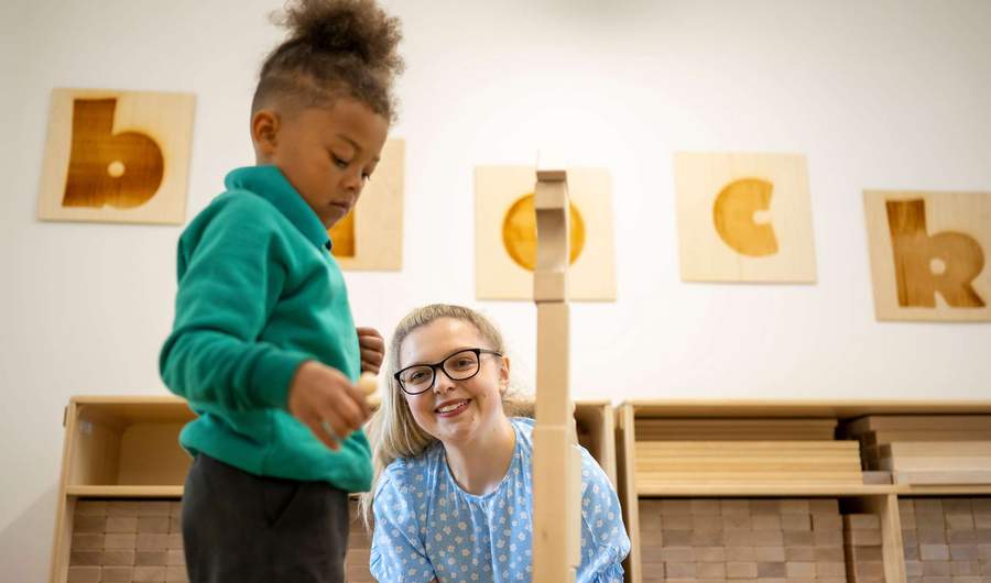 A child plays with wooden blocks as a teacher looks on.