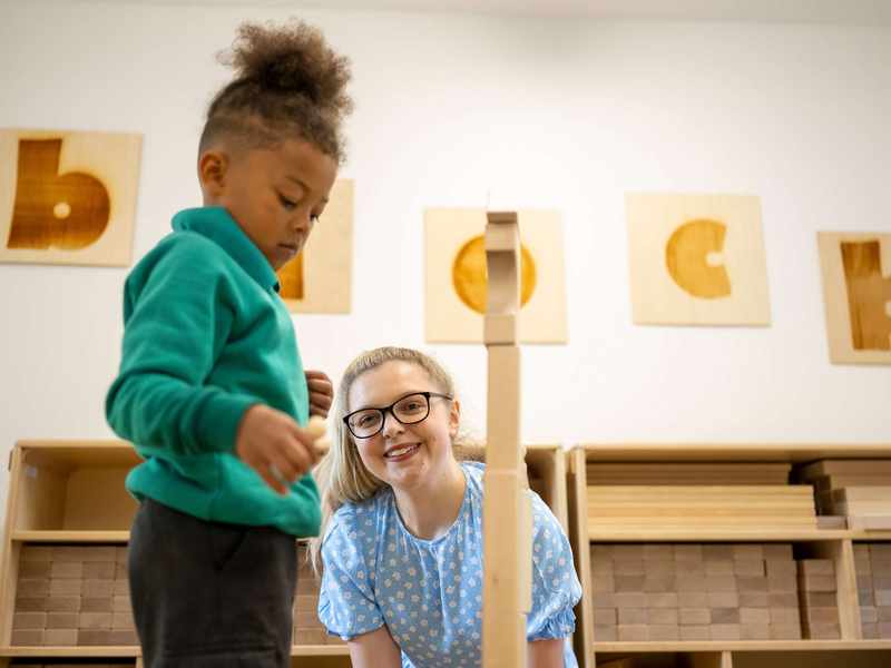 A child plays with wooden blocks as a teacher looks on.