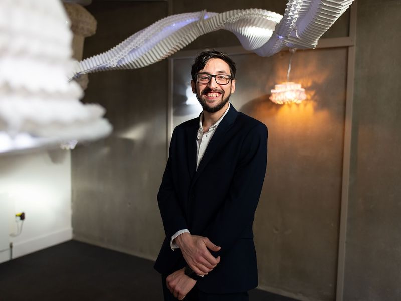 A young adult in a black suite stands underneath an art installation