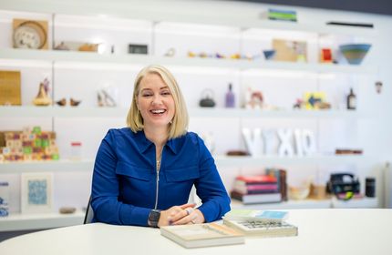 A woman in a blue zipped shirt sits at a desk in front of decorative bookcases