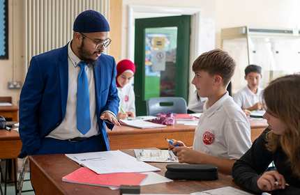 A teacher in blue suit and tie stops to talk with a pupil working at their classroom desk