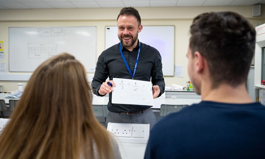A teacher holds up a small handheld whiteboard displaying a sum to two students