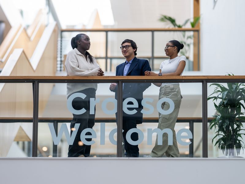 Three students stand talking together on a second floor mezzanine in the Cardiff School of Management building
