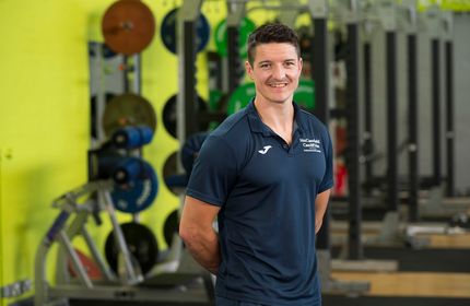 A man in Cardiff Met branded t-shirt stands with arms behind their back in an on-campus gymnasium