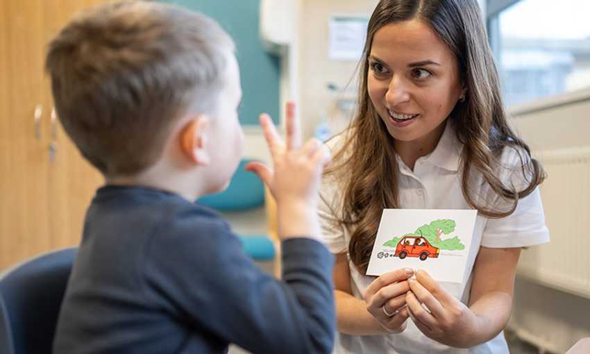 A female speech therapist with long brown hair, wearing a white polo shirt, is engaging with a young child in a therapy session. She is holding a flashcard with an illustration of a red car, while the child gestures with their hands. They are in a well-lit clinical setting with educational materials visible.