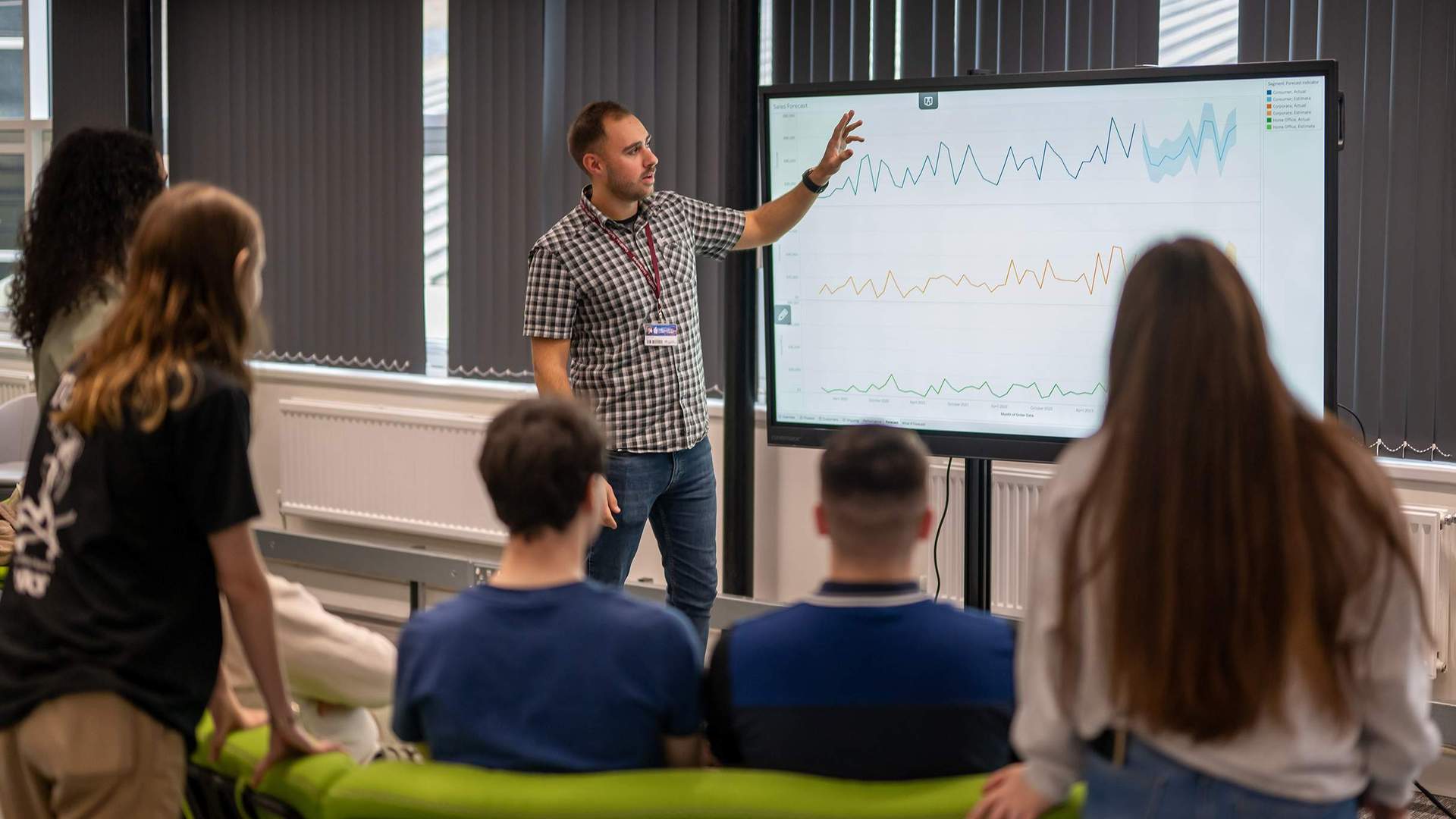 A person stands in front of a small group of people, pointing to charts on a large screen.
