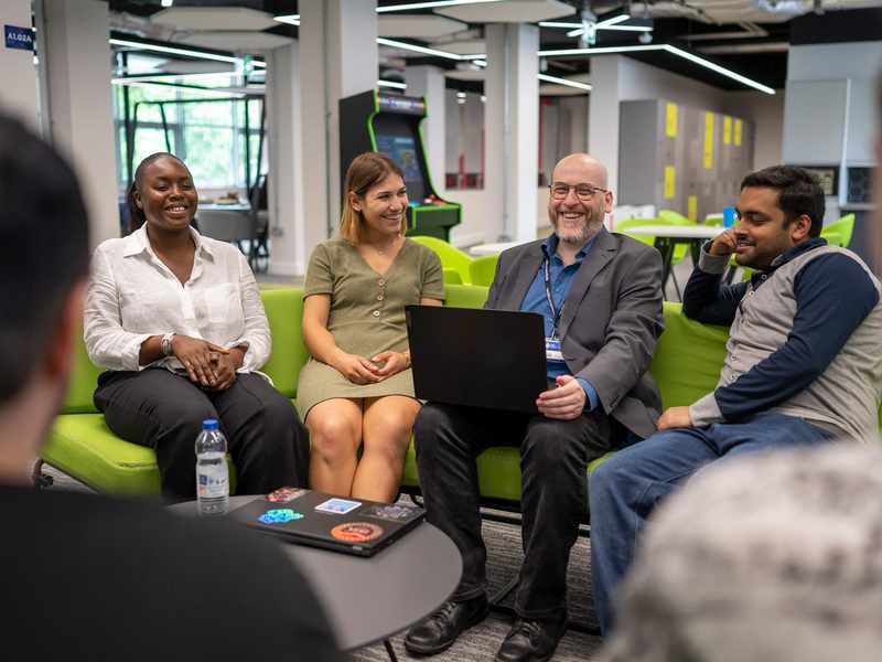 A group of people sit around a small round table. One person has an open laptop computer on their lap.