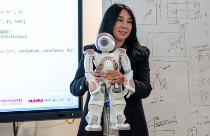 A woman in a black suit holds up a humanoid robot in front of her with both hands