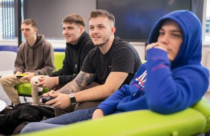Four students sit on a curved green sofa. One is holding a gaming controller and they are all looking in the same direction
