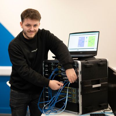 A young adult smiles for the camera as they work on a computer tower and adjust the attached cables