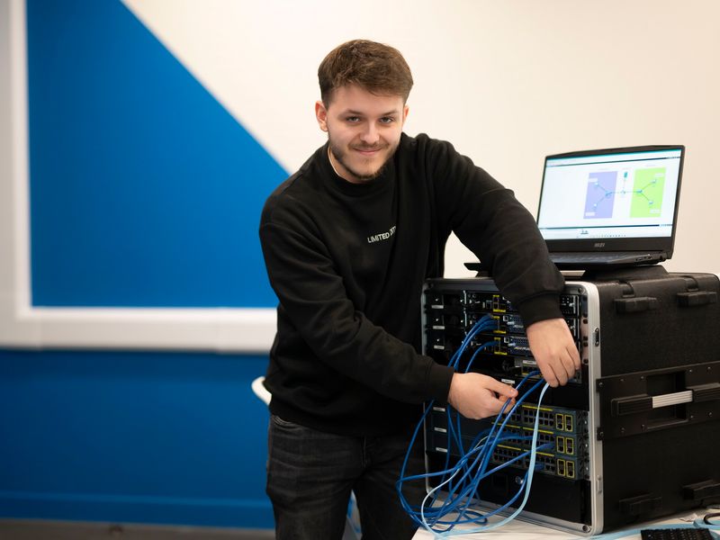 A young adult smiles for the camera as they work on a computer tower and adjust the attached cables