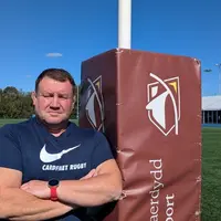 A man stands next to a rugby post, dressed in athletic clothing, with a rugby field visible in the background.
