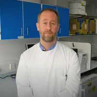 A man wearing a lab coat stands beside a desk, indicating a professional laboratory setting.