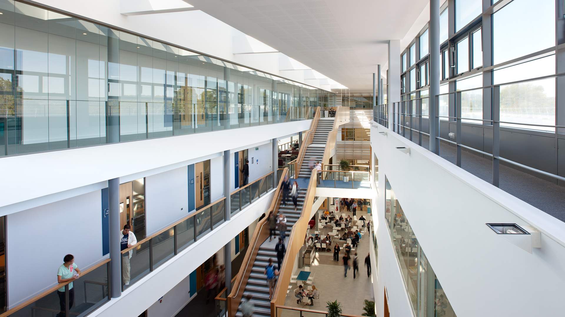 A modern, spacious atrium with a large wooden staircase and multiple people walking. The area has glass railings, plants, and abundant natural light from skylights above. The scene conveys a busy, contemporary office or educational environment.