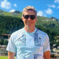 A man wearing sunglasses stands in a field with mountains visible in the background.