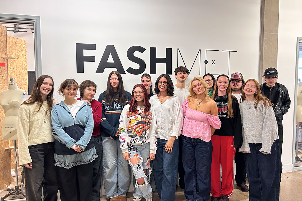 Group of fashion students and an instructor posing in front of a “FASHMET” wall.