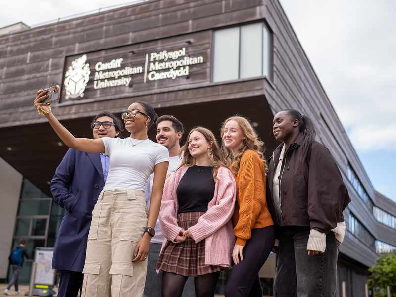 A group of four people walking and talking outdoors, holding coffee cups. Theyre dressed in casual winter clothing, with trees and modern buildings in the background under a partly cloudy sky.