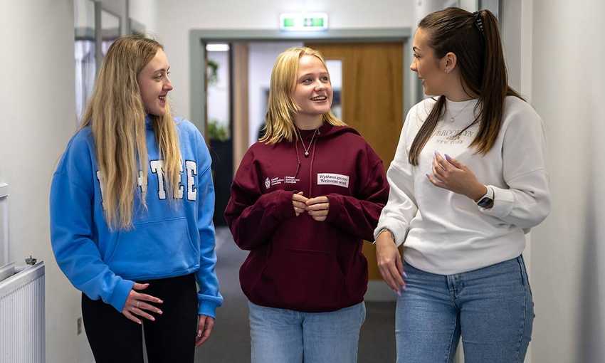 Three young women walk down a hallway together, wearing casual clothes and having a conversation.