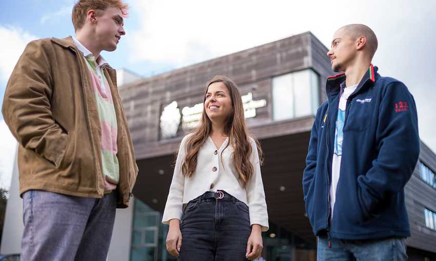Three students stand outside a building, engaged in conversation and enjoying their time together in a campus setting.