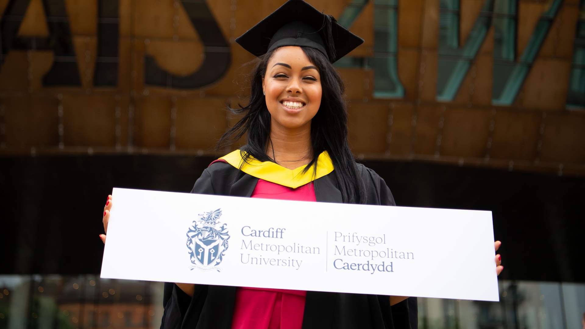 A Cardiff Met student, in graduation cap and gown, stands holding a sign featuring the Cardiff Met logo and name
