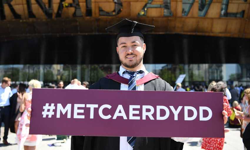A student wearing a graduation cap and gown, stands outside the Wales Millennium Centre holding a #MetCaerdydd branded sign