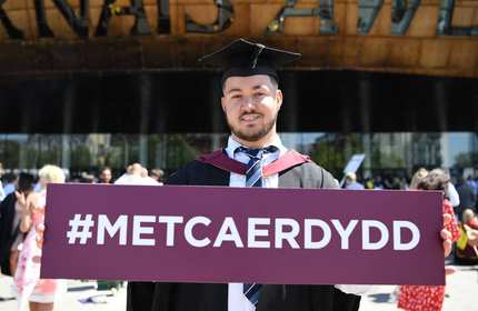 A student wearing a graduation cap and gown, stands outside the Wales Millennium Centre holding a #MetCaerdydd branded sign