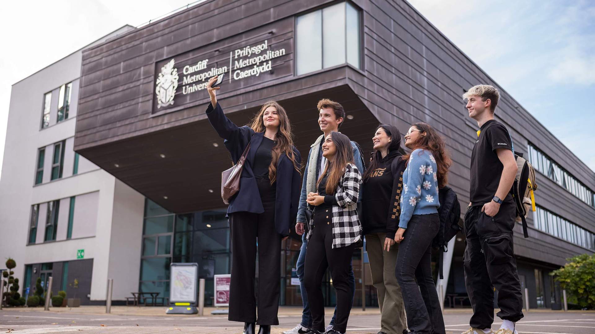 Six young adults stand huddled together outside the Cardiff School of Management as one of them takes a selfie on a mobile phone.