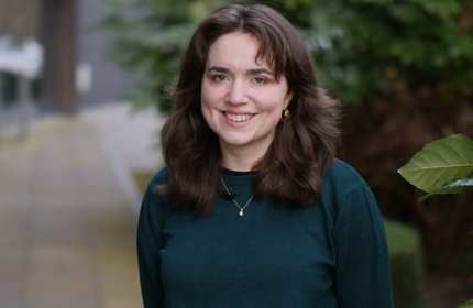 A young woman with long brown hair dressed in a green top, exuding a calm and composed demeanor.