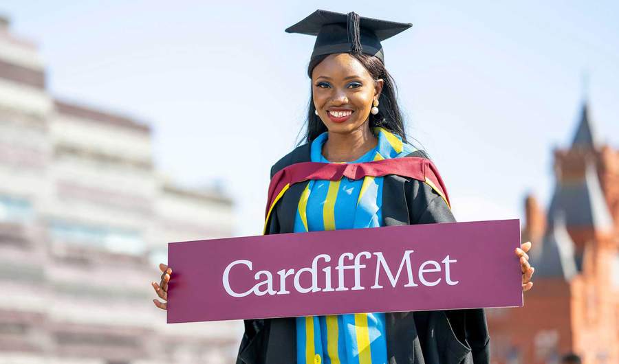 A woman in a graduation gown proudly holds a sign that reads 